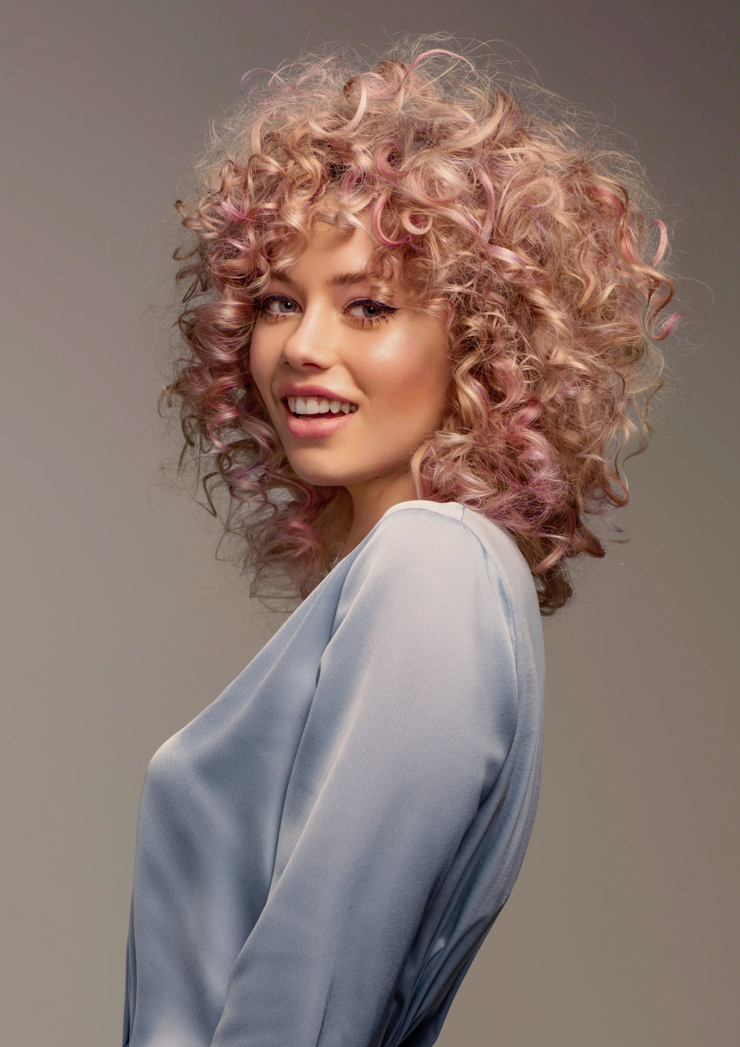 Editorial — woman, voluminous curly pink-blonde hair, blue blouse, grey studio
