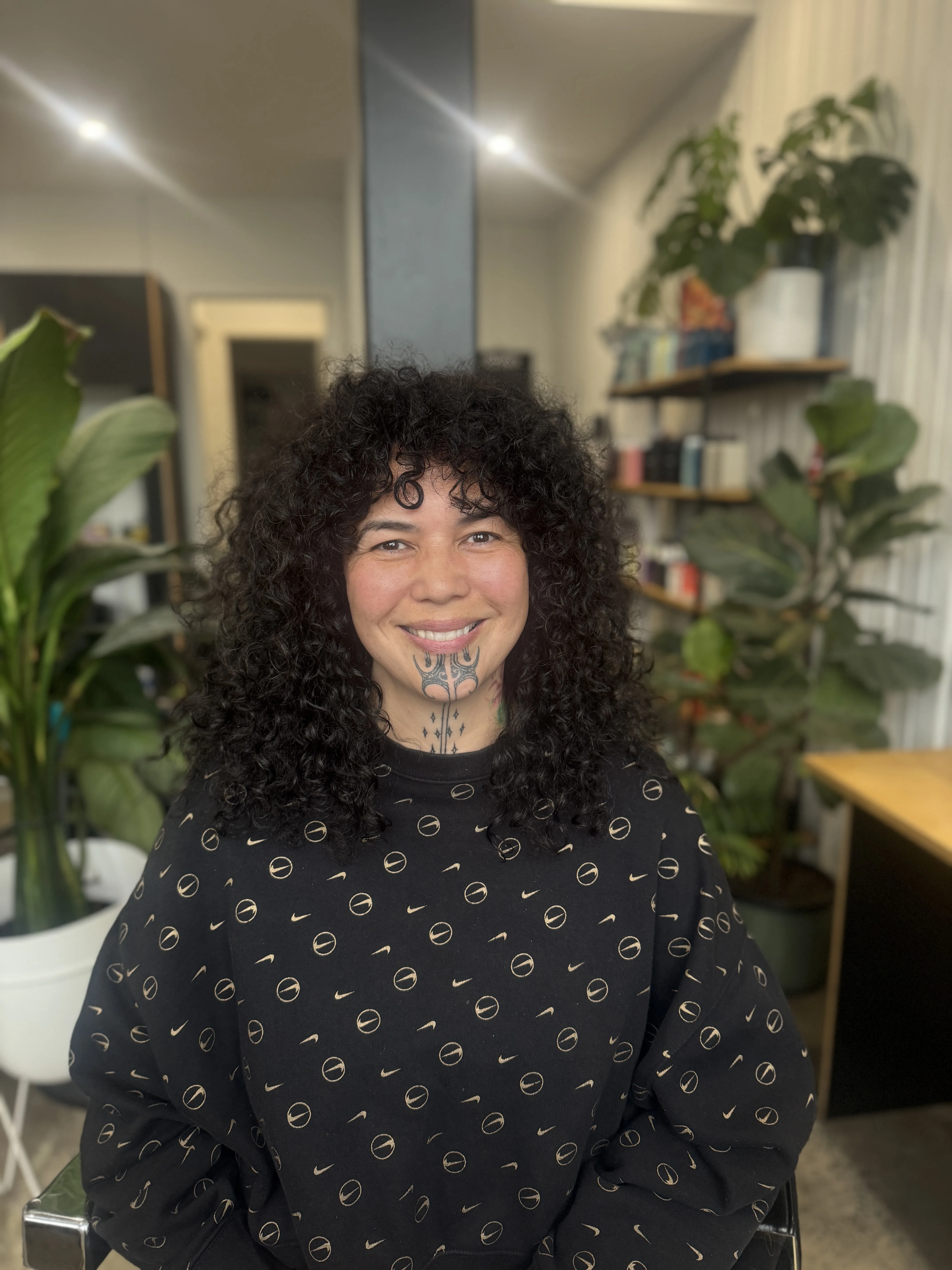 In-salon client — smiling woman, dark curly hair with bangs, chin tattoo, in-salon